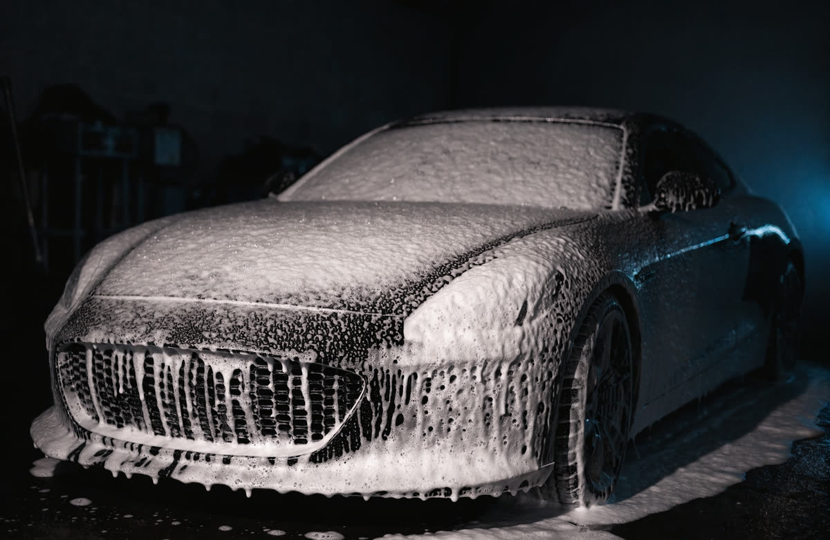 A dark performance car blanketed in thick foam-cannon suds before contact washing