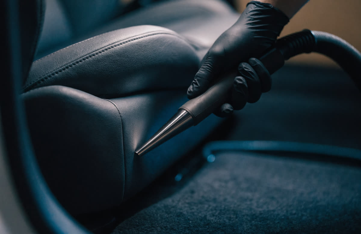 A gloved hand guiding a vacuum crevice tool along the seam of a car seat